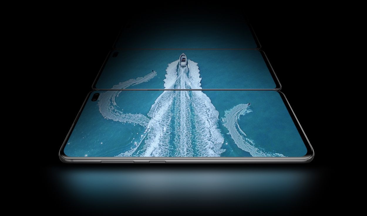 Trois téléphones Galaxy S10 plus en mode paysage, posés à plat au-dessus de celui qui le précède, vus sous un léger angle depuis le côté du bouton Bixby and Volume.  Sur les trois écrans, il y a une vue aérienne d'un bateau dans l'eau avec deux skieurs nautiques, montrant l'étendue de l'écran Infinity-O Display.